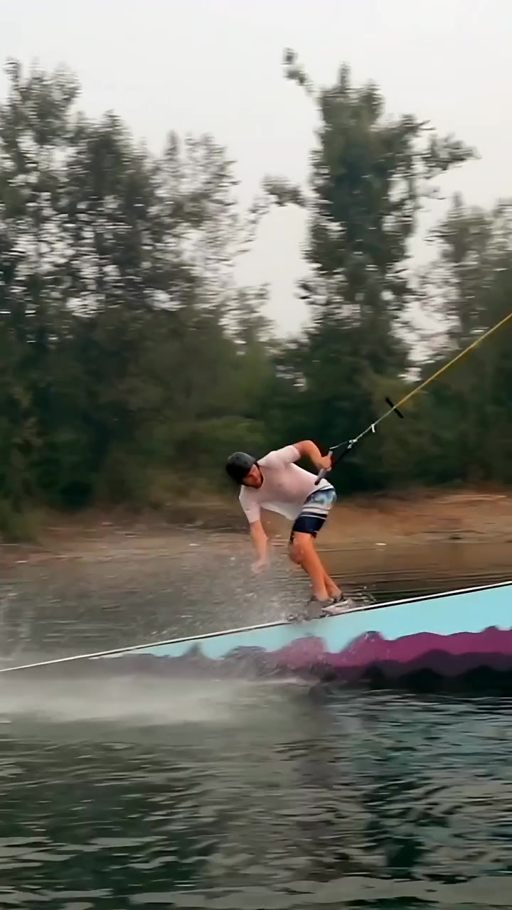 Video verticale: Cavo di traino che tira un rider su wakeboard, con casco e pantaloncini, su un lago che scivola su un rail per divertimento.