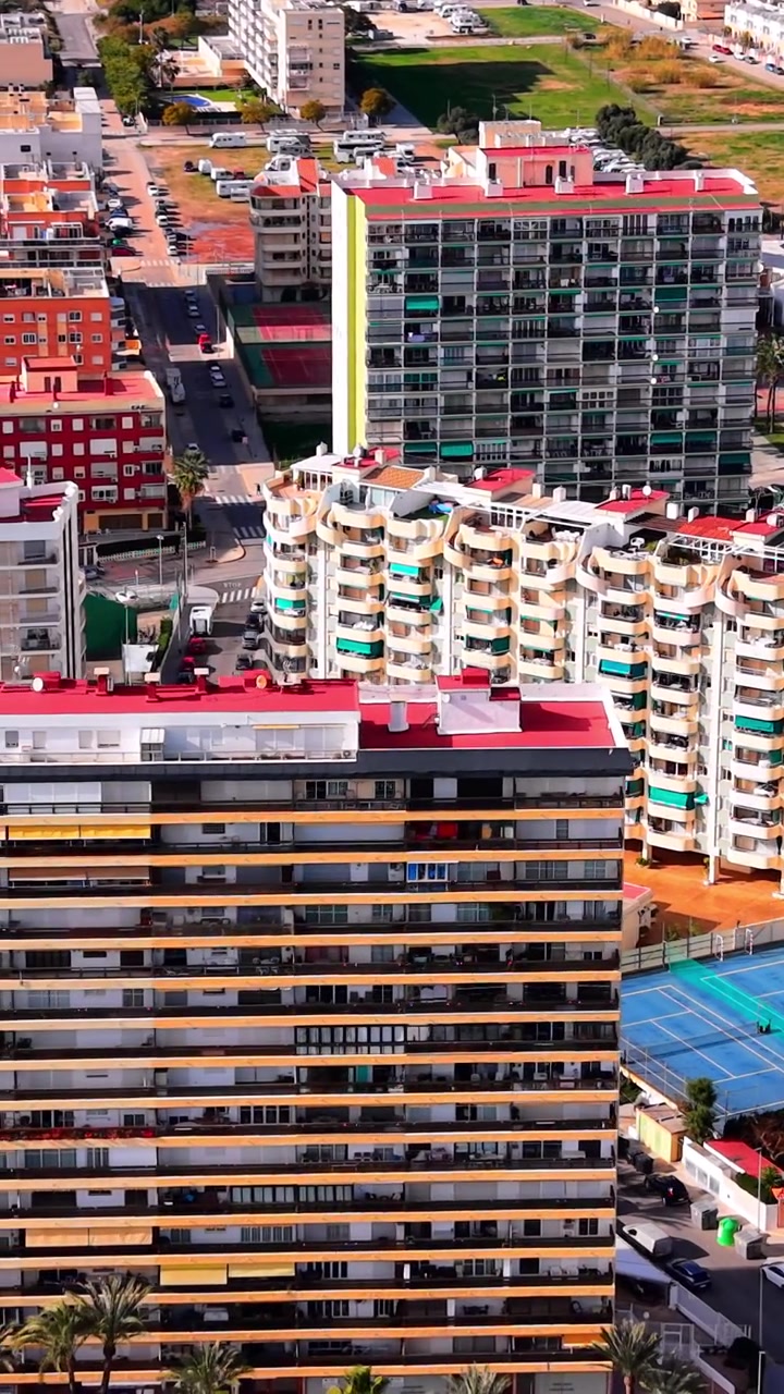 Edifícios costeiros e quadra de tênis azul. Vista elevada, arquitetura de resort, telhados de telhas vermelhas, rua urbana, dia claro e ensolarado, mercado imobiliário mediterrâneo. Vídeo vertical