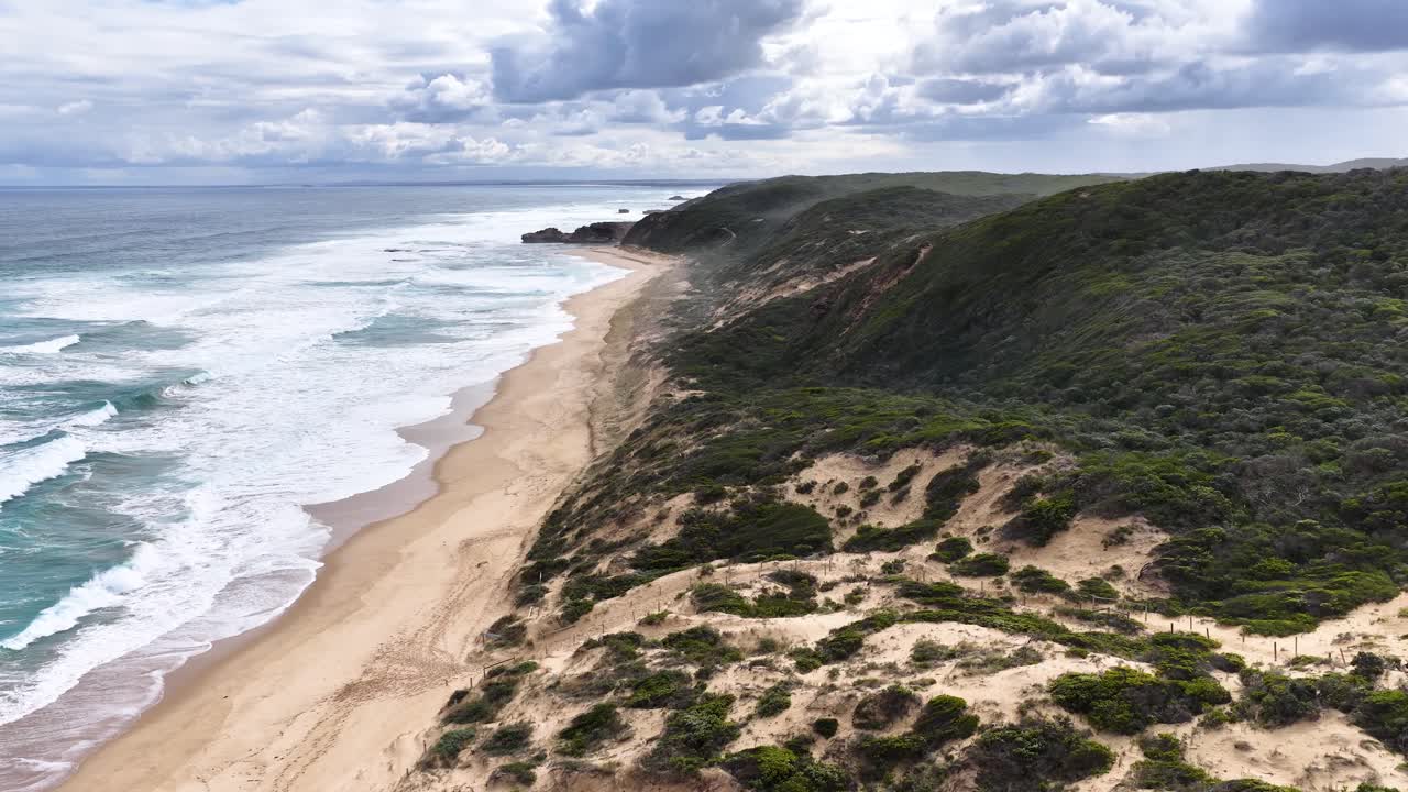 Drone glides above sandy cliffs, green vegetation, and crashing waves under dramatic cloudy skies