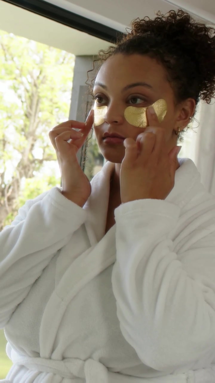 Verticale video: Houdt gouden patches vast, vrouw drukt deze onder haar ogen in een witte badjas thuis voor huidverzorging.