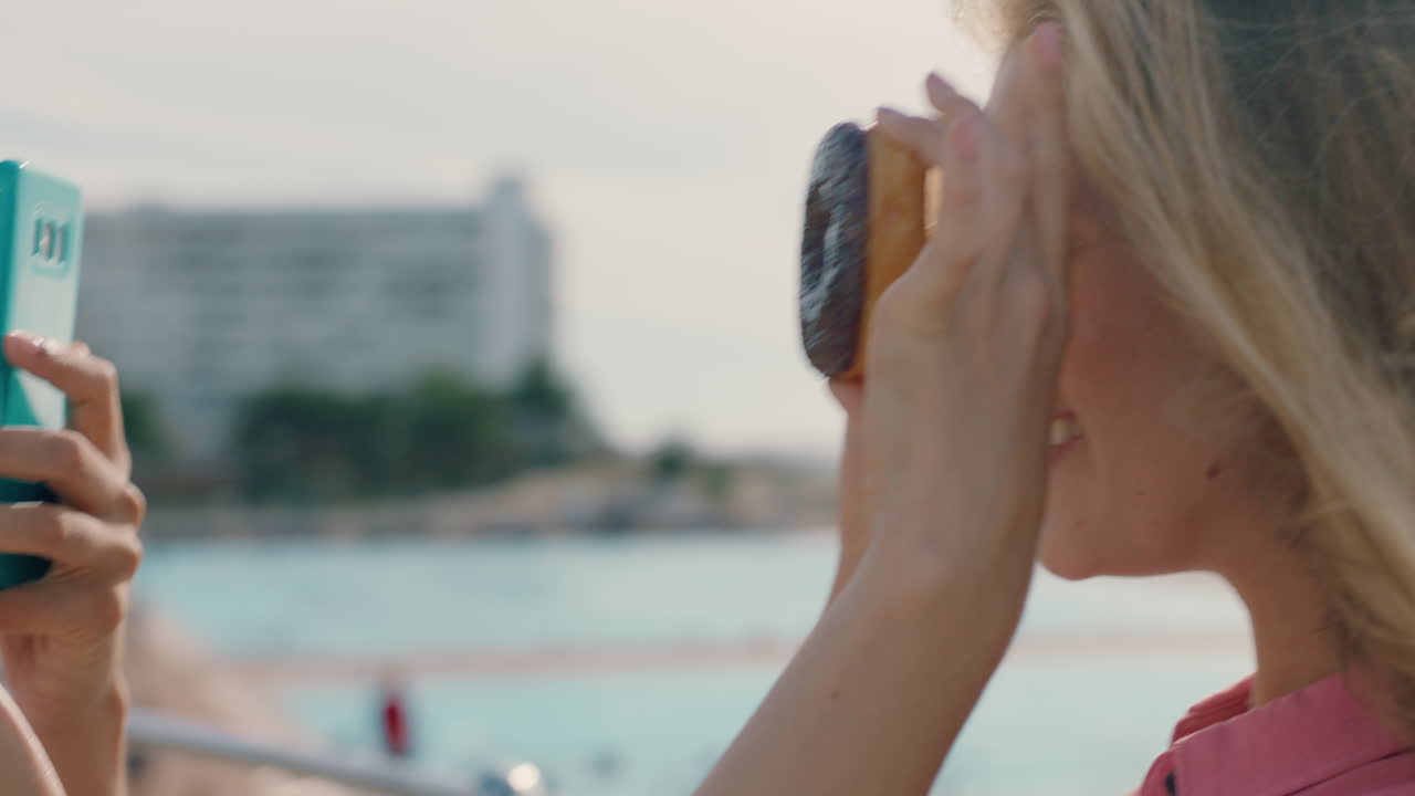 bella donna che posa con le ciambelle sulla spiaggia migliore amica che scatta foto usando lo smartphone condividendo weekend al mare sui social media godendosi il divertimento estivo 4k