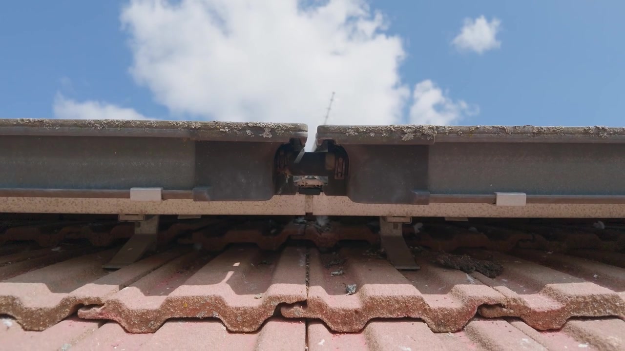 Solcellepaneler på taket panorerer ned fra blå himmel og skyer til røde takstein, TV-antenne og fjær under panelene, noe som antyder at fugler hekker under panelene på et hus.