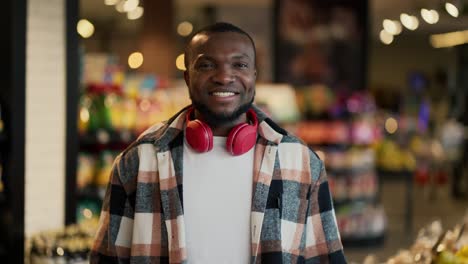 Portrait-of-a-happy-young-man-with-Black-skin-color-in-a-plaid-shirt-and-red-wireless-headphones-who-is-smiling-and-posing-near-the-counters-in-a-modern-grocery-store