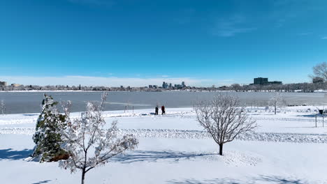 drone moving past snowy trees at sloan lake during winter storm, revealing a family walking with denver city in the background