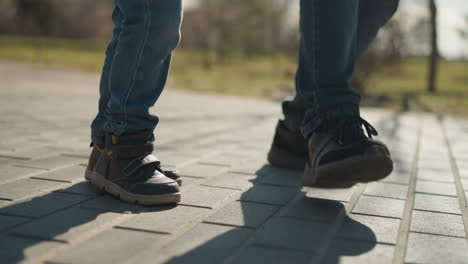 a close-up of two people s legs, one adult and one child, as they stamp their right leg together on a paved path, both are wearing blue jeans and sturdy boots