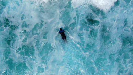 AERIAL-:-Birdseye-view-of-surfer-battling-the-waves,-current-and-white-wash-in-a-turbulent-ocean