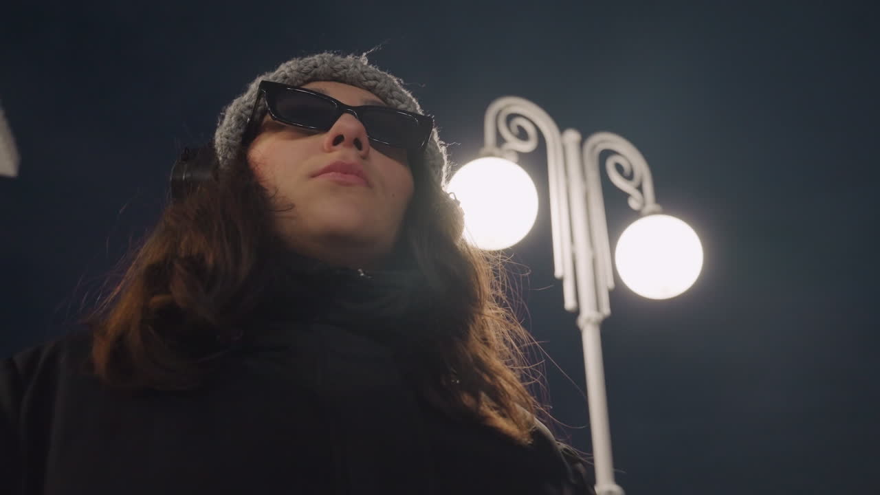 Bajo la farola, una mujer se quita las gafas de sol. Planos cinematográficos en picado capturan una mirada segura y el ritmo de los auriculares, halo de luz de la calle y bokeh, abrigo de invierno elegante y energía de modelo serena, gestos de baile lentos.
