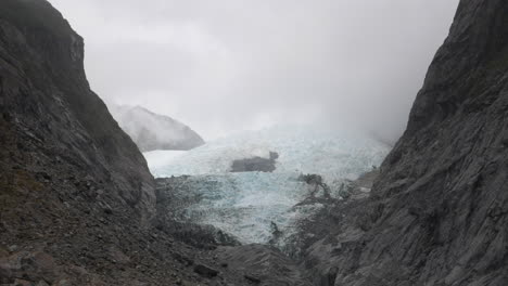 rising shot of the frozen rob roy glacier, new zealand, surrounded by grey rocks, with fog moving above, on a cloudy day