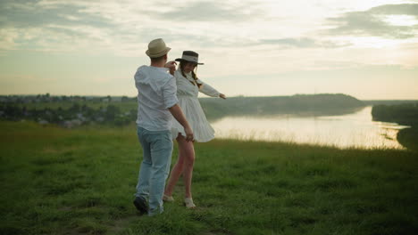 a man spins his wife around on a grassy hill beside a peaceful lake at sunset. the man is dressed in a white shirt, hat, and jeans, while the woman wears a white dress and black hat, a romantic scene