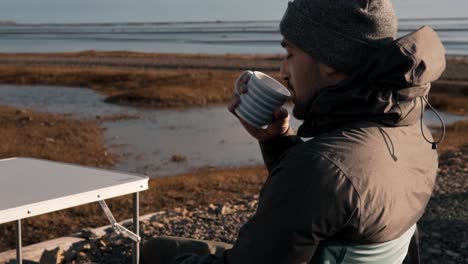 A warmly dressed traveler sips coffee from a mug at a portable table, taking in the serene beauty of a Stokksnes and Vestrahorn sunrise