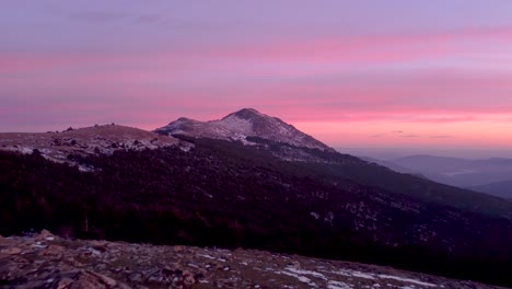 Aerial-view-flying-forward-with-mountaineer-in-red-coat-standing-on-a-ridge-during-colorful-sunset-in-winter-with-snow-on-mountain-peaks-in-Madrid,-Spain