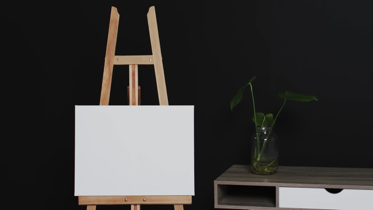 Video of white canvas sign on wooden easel with copy space and plant on desk on black background