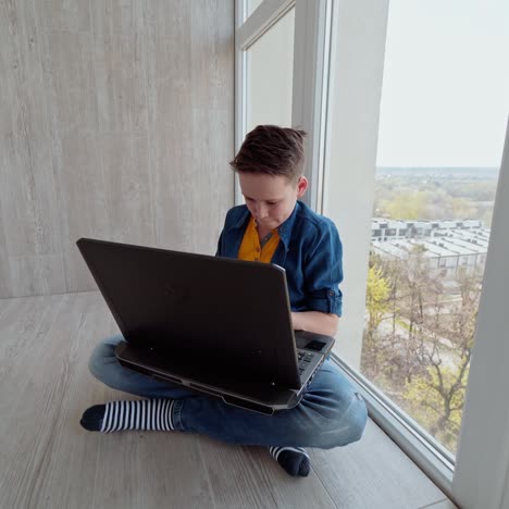 teen boy plays on the gadget on a balcony. boy sitting by the window and playing video games on a modern laptop expresses different emotions.