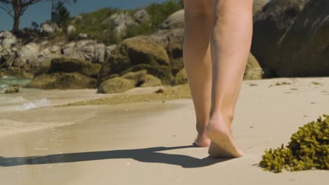 A-close-up-shot-tracking-behind-a-female-walking-along-an-ocean-beach,-tilting-up-to-reveal-the-surrounding-rocky-landscape-and-the-intended-destination