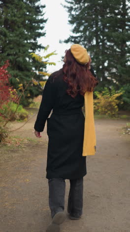 vista trasera de una mujer joven con cabello rojo ondulado largo con abrigo negro, boina amarilla y pañuelo a juego caminando por un camino de tierra a través de un exuberante parque verde con colorido follaje de otoño, mirando a su alrededor