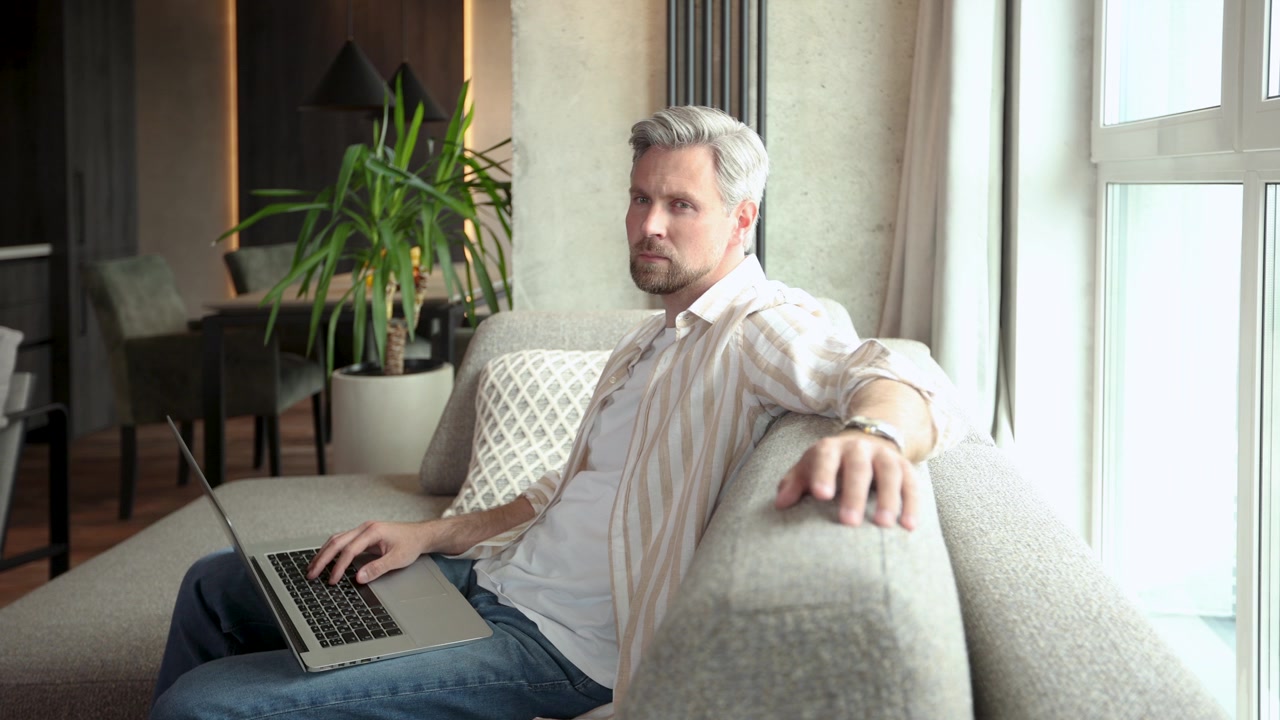 Man Working on Laptop on Sofa at Home Looks at Camera