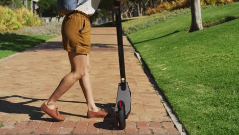 mujer afroamericana con auriculares y mochila montando un scooter en el parque