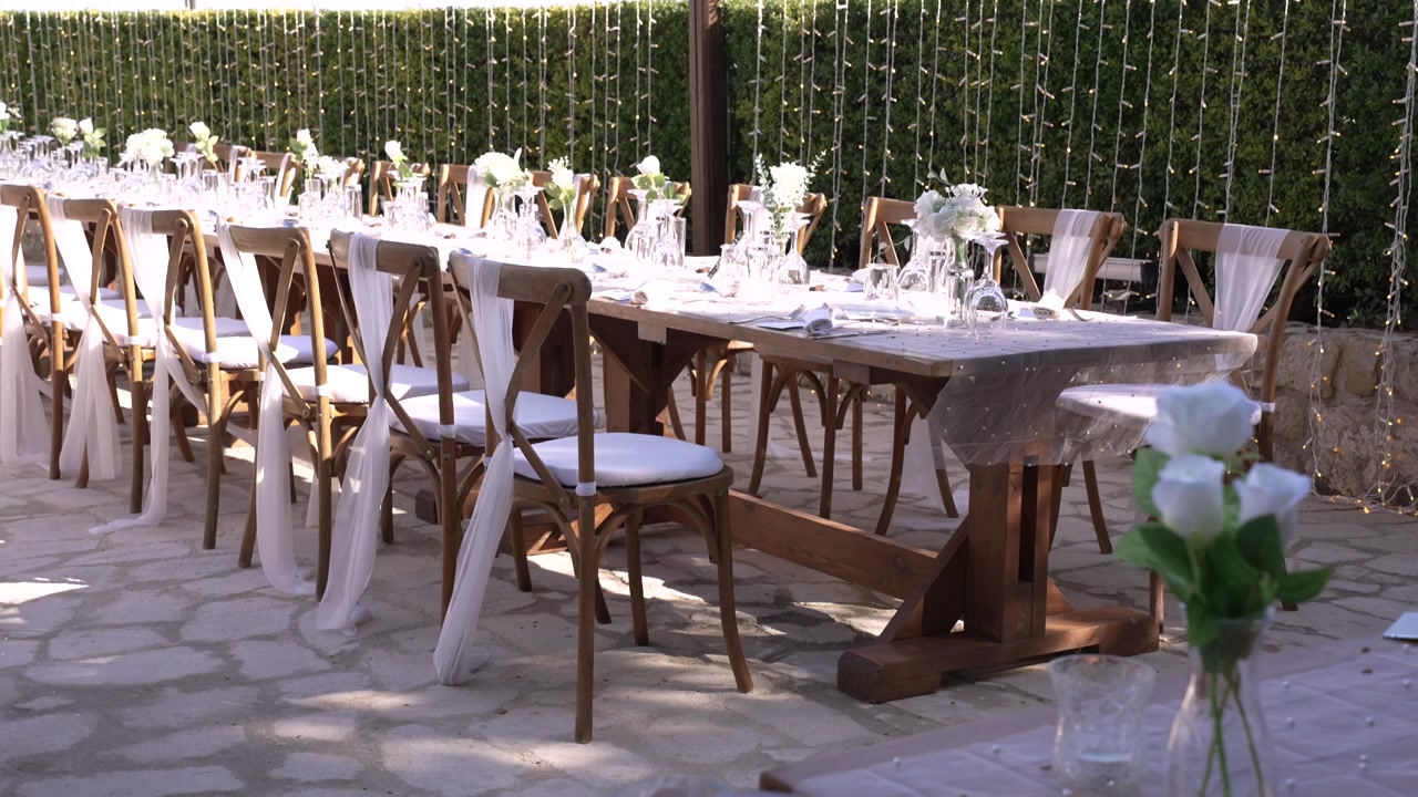 Tables are arranged in an outdoor garden setting. Each table has white flowers in vases. The space is ready for a gathering with decorations, and lights are hanging above.