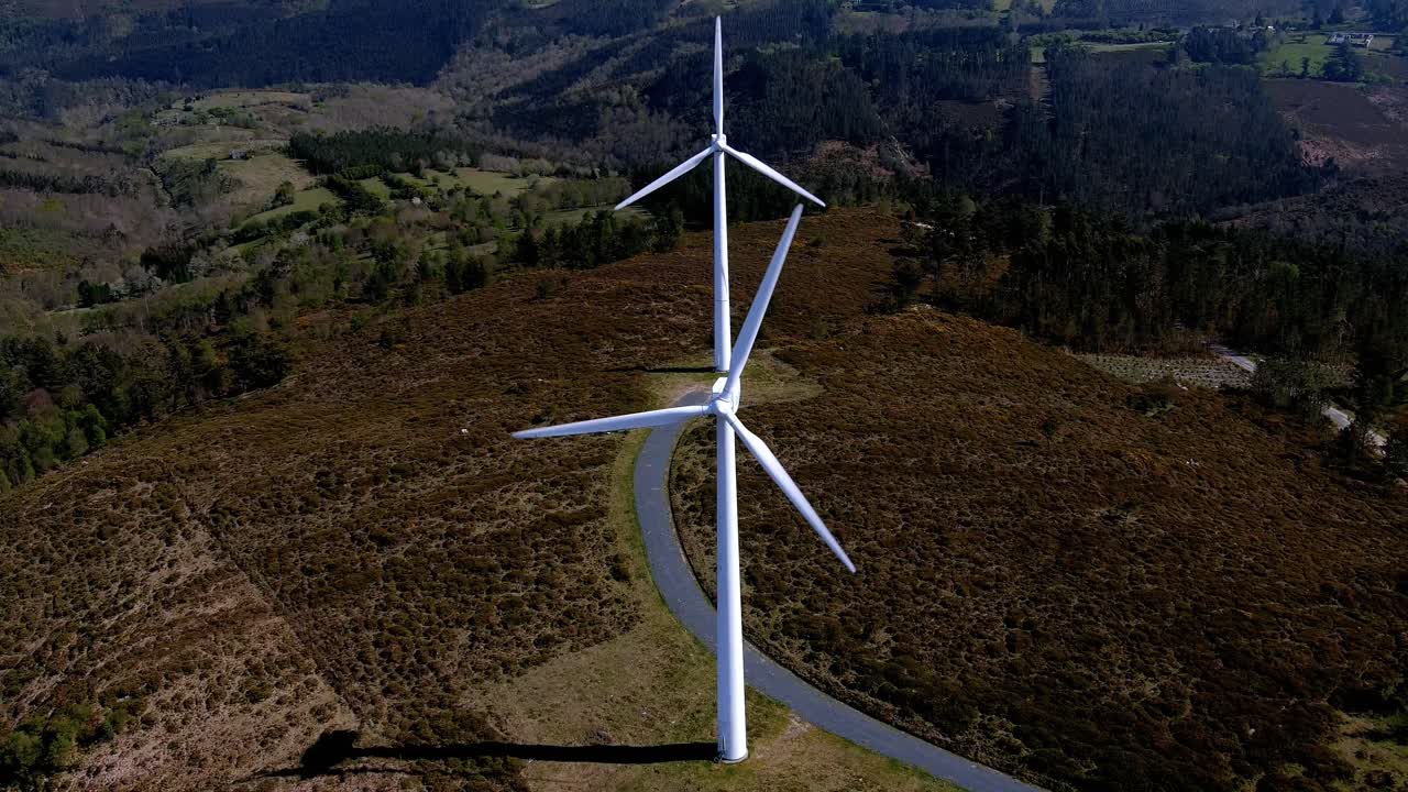 dos turbinas eólicas alineadas girando sus palas en las montañas con pequeños bosques verdes de árboles en una tarde soleada de cielo azul