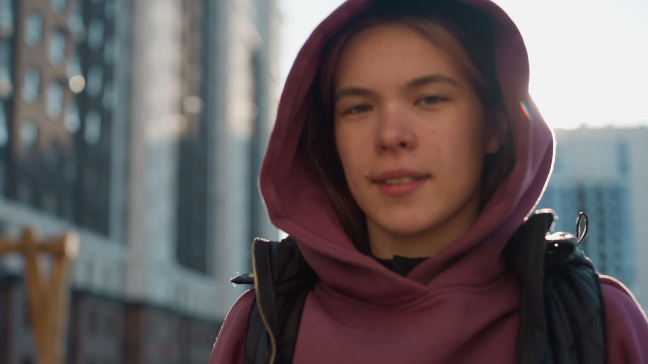 Portrait of young fair athlete smiling under sunny sky in urban park setting with confident calm expression and relaxed posture, hoodie vest combo and sunlit highlights in vibrant city scene