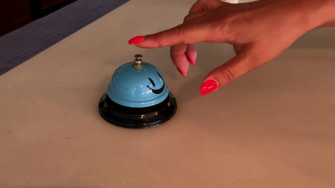 Descripción: Mano con uñas rojas extendiéndose hacia una campana de servicio azul con cara sonriente en un mostrador de recepción.