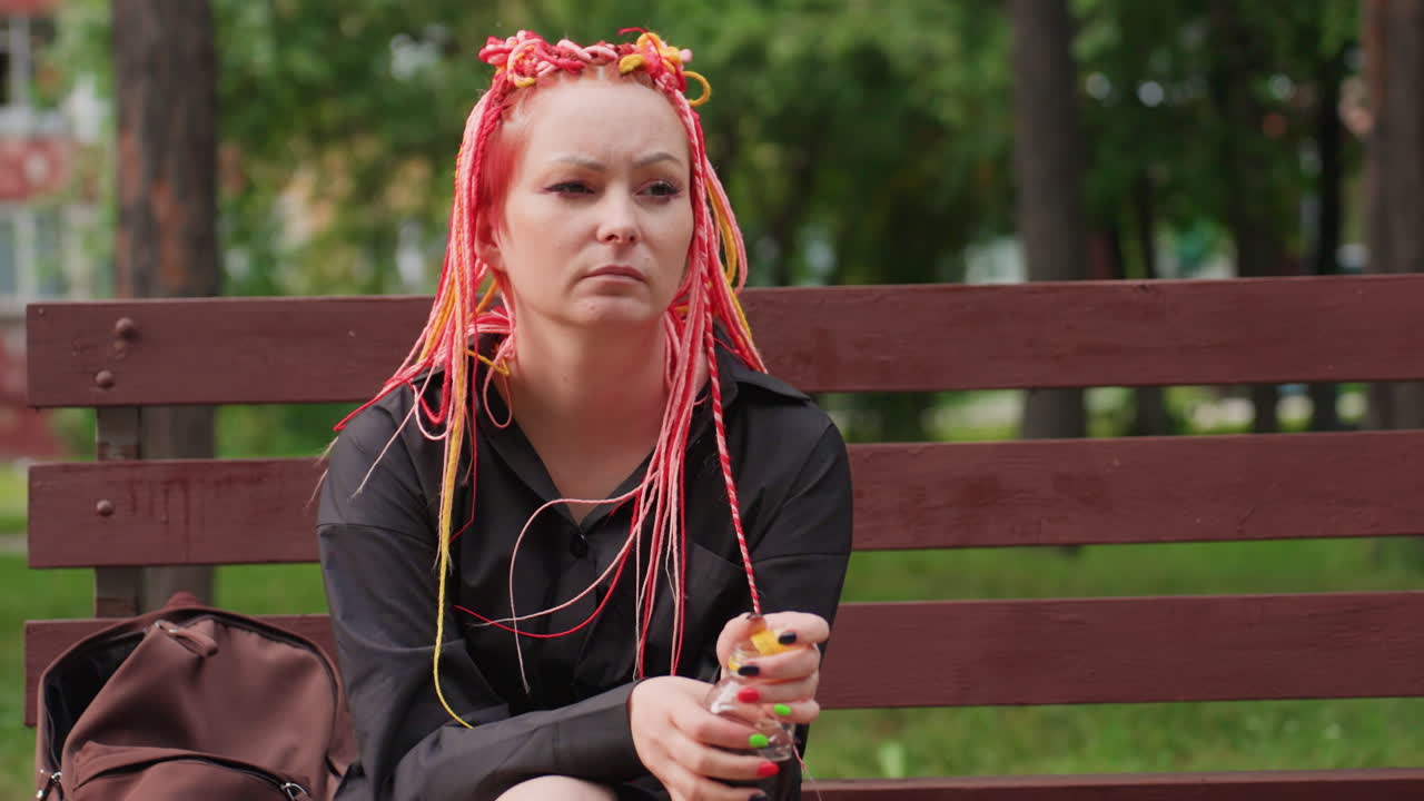 나무 벤치에 앉아 똑바로 앉아 병을 들고 있는 백인 여성의 넓은 미디엄 샷, 침착하고 금욕적인 자세, 화려한 땋은 머리, 주변 공원의 나무와 비어 있는 산책로는 인내와 회복력을 암시합니다.