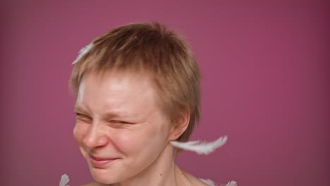 a woman with short blonde hair smiles and laughs on a pink background, with white feathers falling around her
