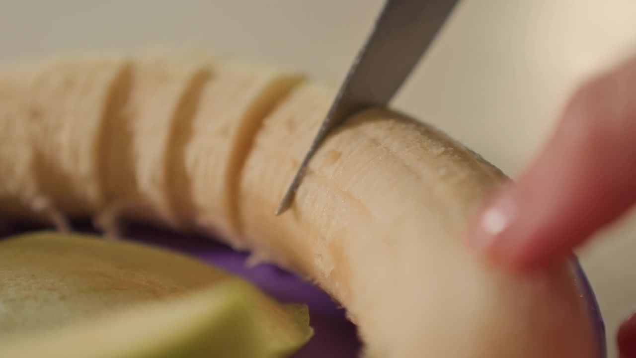 Macro kitchen scene showing knife cutting banana into short diagonal pieces while fingertip steadies curved fruit on plate rim, with pale flesh exposed, peel detail, soft light, precise motion