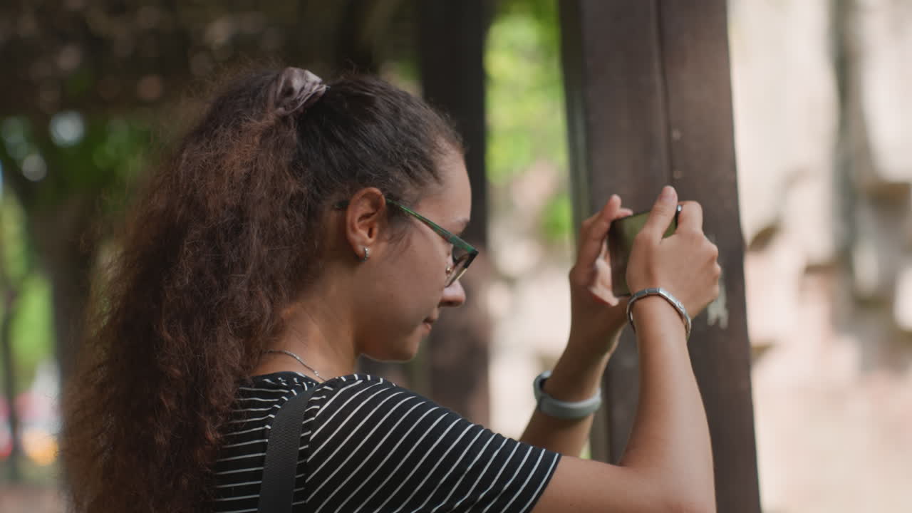 Young woman in striped top composing closeup on smartphone by wooden post, concentrated and steady hands casual content creator and student roles, soft background bokeh, leafy city corner