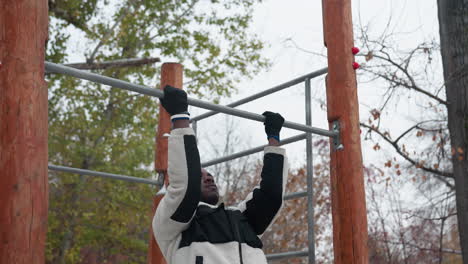 boy performing pull-up exercise on outdoor workout bar in winter. wearing warm fleece jacket and gloves, he grips bar with determination, snow-covered ground, leafless trees, and urban background