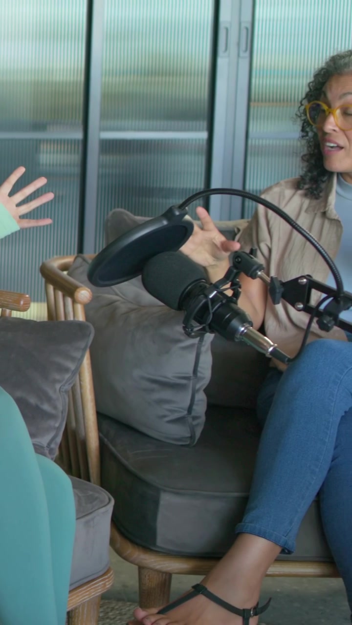Vertikal video: Kamera trækker sig tilbage, kvinde i lyserød top taler til podcast med mikrofon, kopper og plads til tekst.
