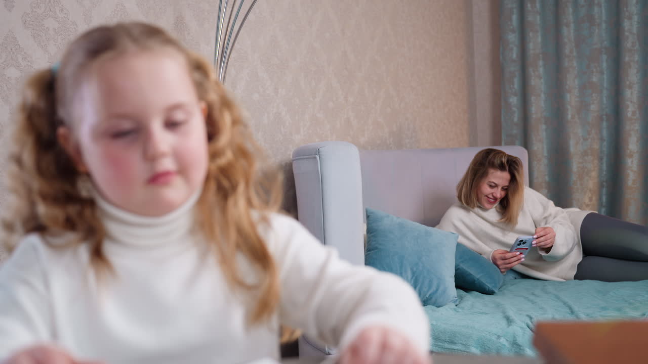 Ребенок сидит за столом и тянется к карандашам, а мать на заднем плане отдыхает на кровати и улыбается, глядя в смартфон. Уютная домашняя обстановка, демонстрирующая детские занятия, семейный образ жизни и многозадачность