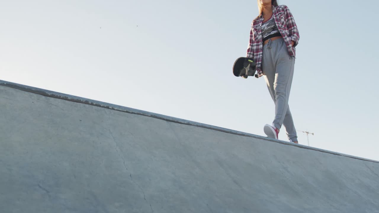 mujer caucásica sonriente, caminando, sosteniendo una patineta en un skatepark