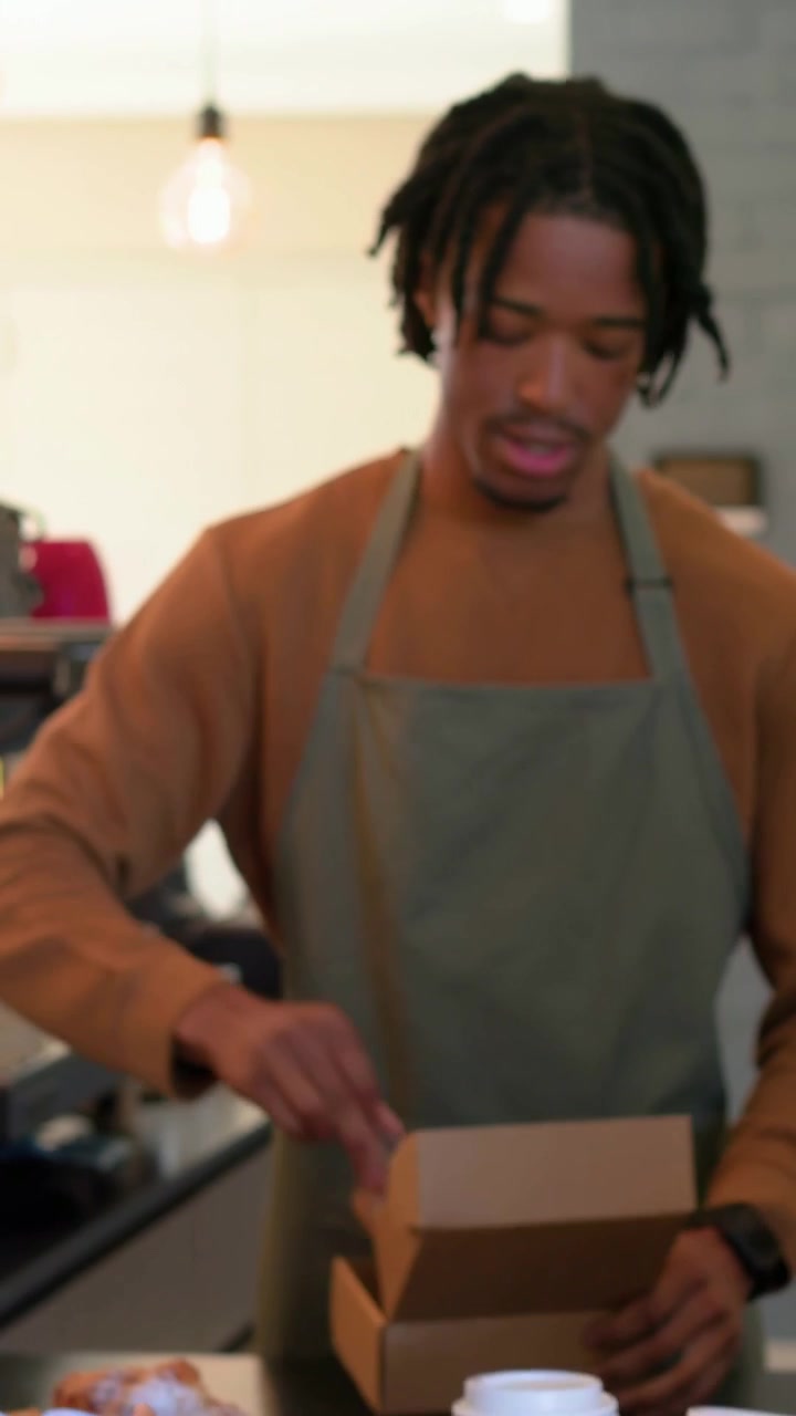 Vertical video: Serving customer at small cafe barista brown sweater green apron packing croissant