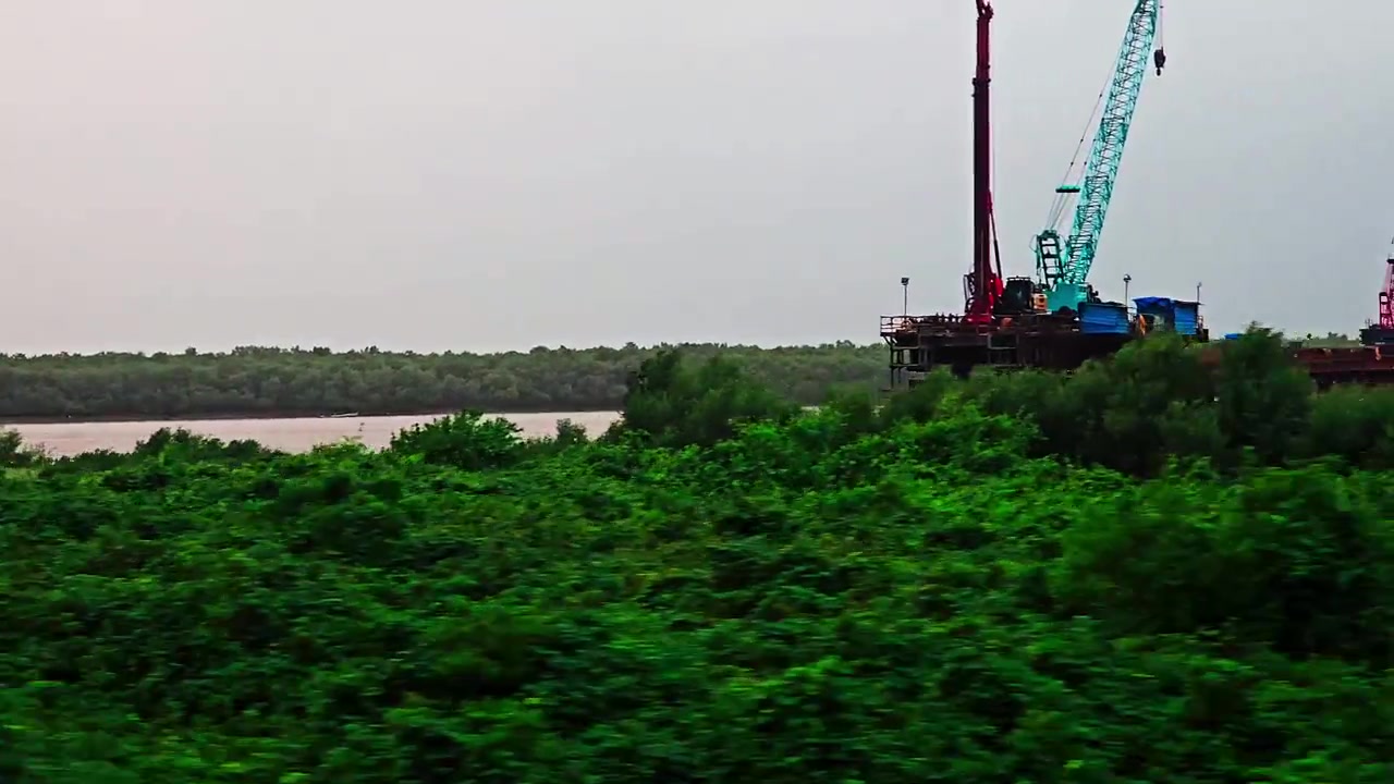 Hindistan'da bir su yolu mavnasında bulunan ağır inşaat vinçlerini ve ekipmanlarını gösteren, yoğun yeşil bitki örtüsünün yanından hızla geçen bir trenden hareket bulanıklığı olan pencere görüntüsü.