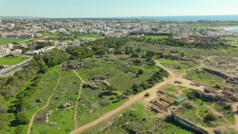 Aerial-ruins-scattered-across-a-green-landscape-in-the-foreground-and-the-modern-cityscape-of-Paphos,-Cyprus,-in-the-distance