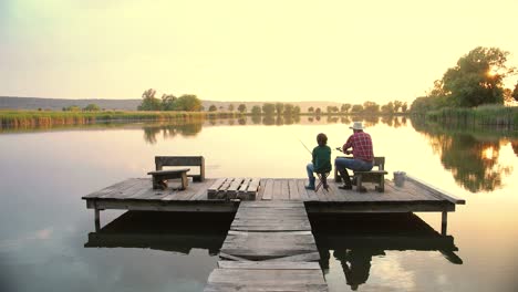 Vista-Trasera-De-Un-Adolescente-Sentado-Con-Su-Abuelo-En-El-Muelle-Del-Lago,-Hablando-Y-Pescando-Juntos