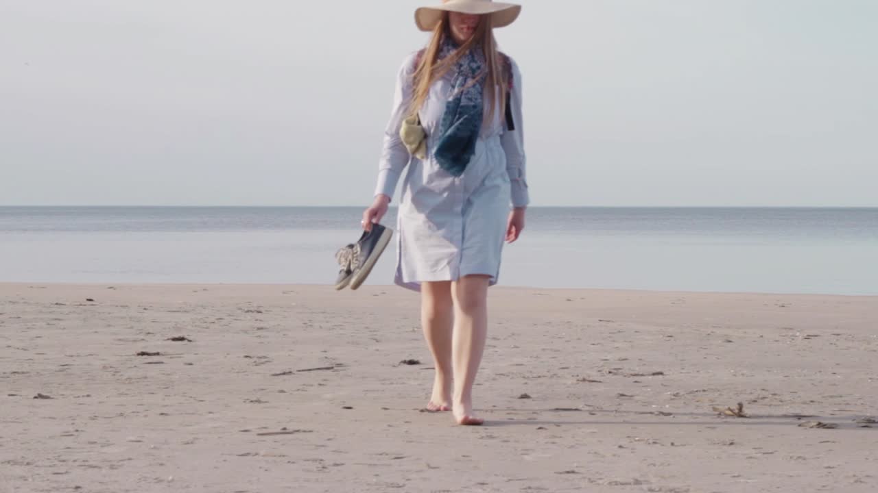바다에서 카메라를 향해 걸어가는 백인 여성은 여름옷과 모자를 쓴다