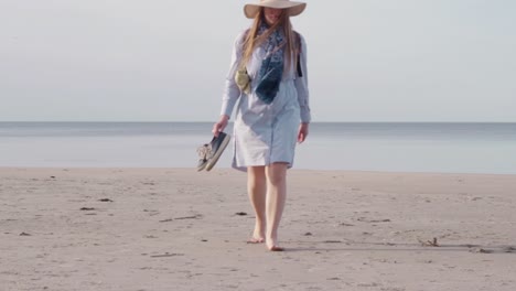 caucasian woman walking towards camera at sea wears summery clothes and hat