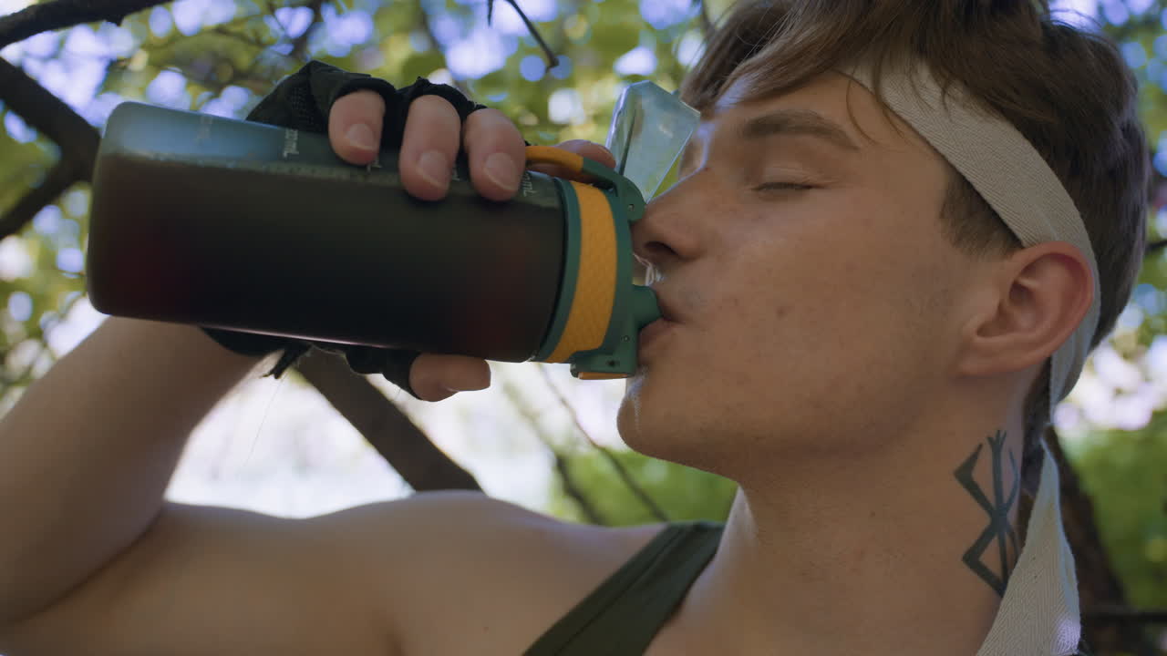 young man wearing headband and tank top pausing training to drink coffee from water bottle under leafy canopy with eyes closed and muscles relaxed amid dappled sunlight creating serene outdoor moment