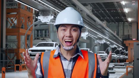 안전 헬을 입은 아시아 남성 엔지니어가 카메라에 미소 짓고 ev 자동차 공장에서 서서 와우라고 말합니다.