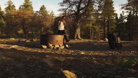 Man-carries-logs-to-chopping-block-in-sunny-forest,-long-shot