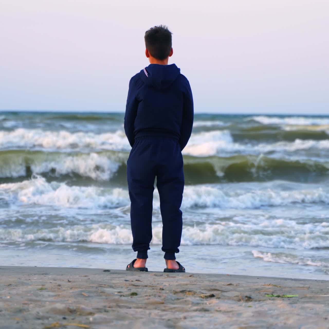 Teenage boy looking at the sea. Backside view of a child in clothes standing on a sandy beach near the sea and looking on waves in summer evening.