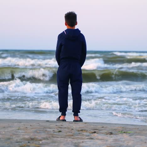 teenage boy looking at the sea. backside view of a child in clothes standing on a sandy beach near the sea and looking on waves in summer evening.