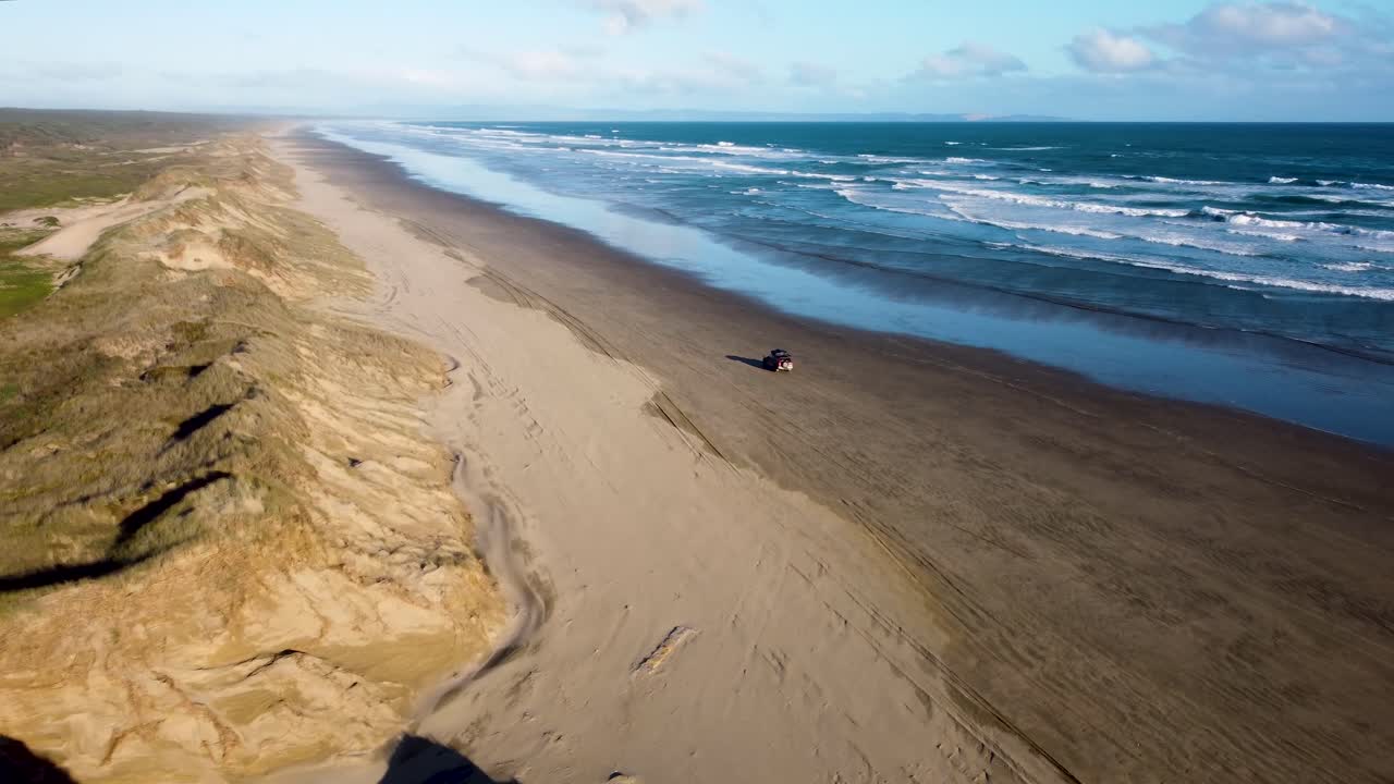 Drone view of offroad 4 wheel drive red car with rooftop tent driving on sand next to the ocean on a long beach on a sunny day at 90 Miles Beach, Northland, New Zealand.