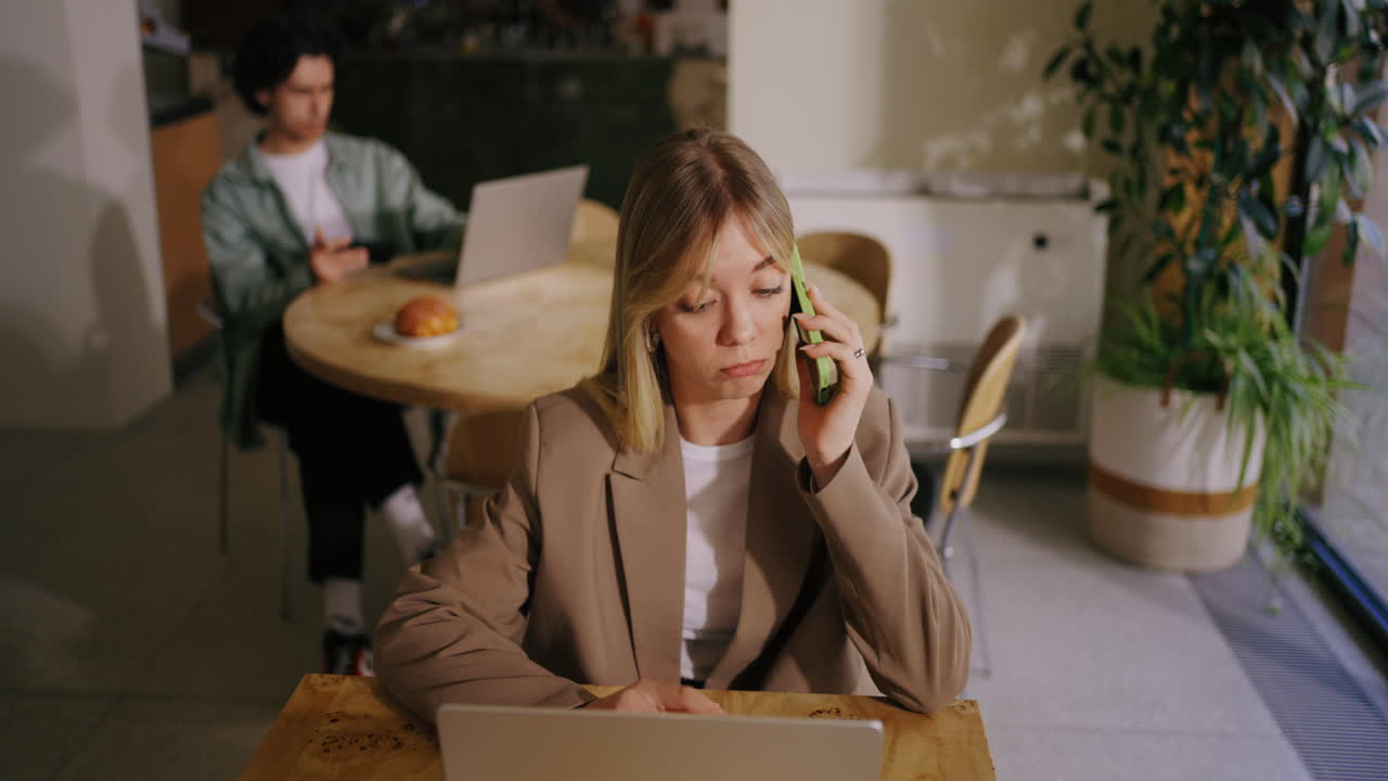 젊은 사업가 여성이 커피에서 노트북으로 일하면서 전화로 얘기하고 있습니다.