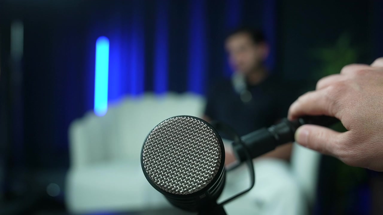 Primo piano di un microfono professionale in uno studio di podcast con l'host che regola l'asta. Ospite sfocato seduto sul divano sullo sfondo, concetto di trasmissione e registrazione multimediale