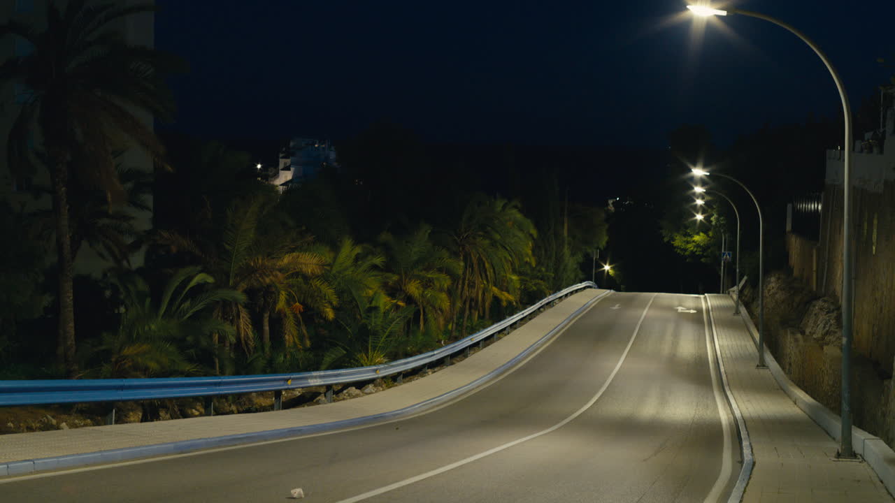 Strada costiera vuota in Spagna in una notte di tempesta, lampioni che brillano mentre le palme si piegano drammaticamente nel forte vento sotto un cielo blu scuro.