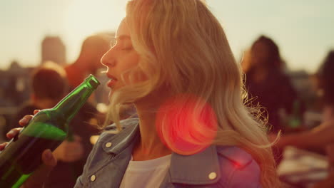 Young-woman-drinking-beer-at-disco.-Smiling-girl-dancing-at-sunset-roof-party.