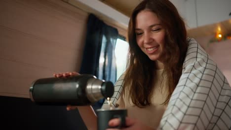 a happy brunette girl wrapped in a white blanket pours herself hot tea into a black mug from a thermos while sitting in a trailer during her picnic in the camp and enjoying the smell and warmth of a hot drink